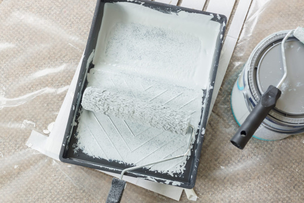 Painting and decorating with rollers and tray filled with light grey paint on a plastic dust sheet with cans in the background